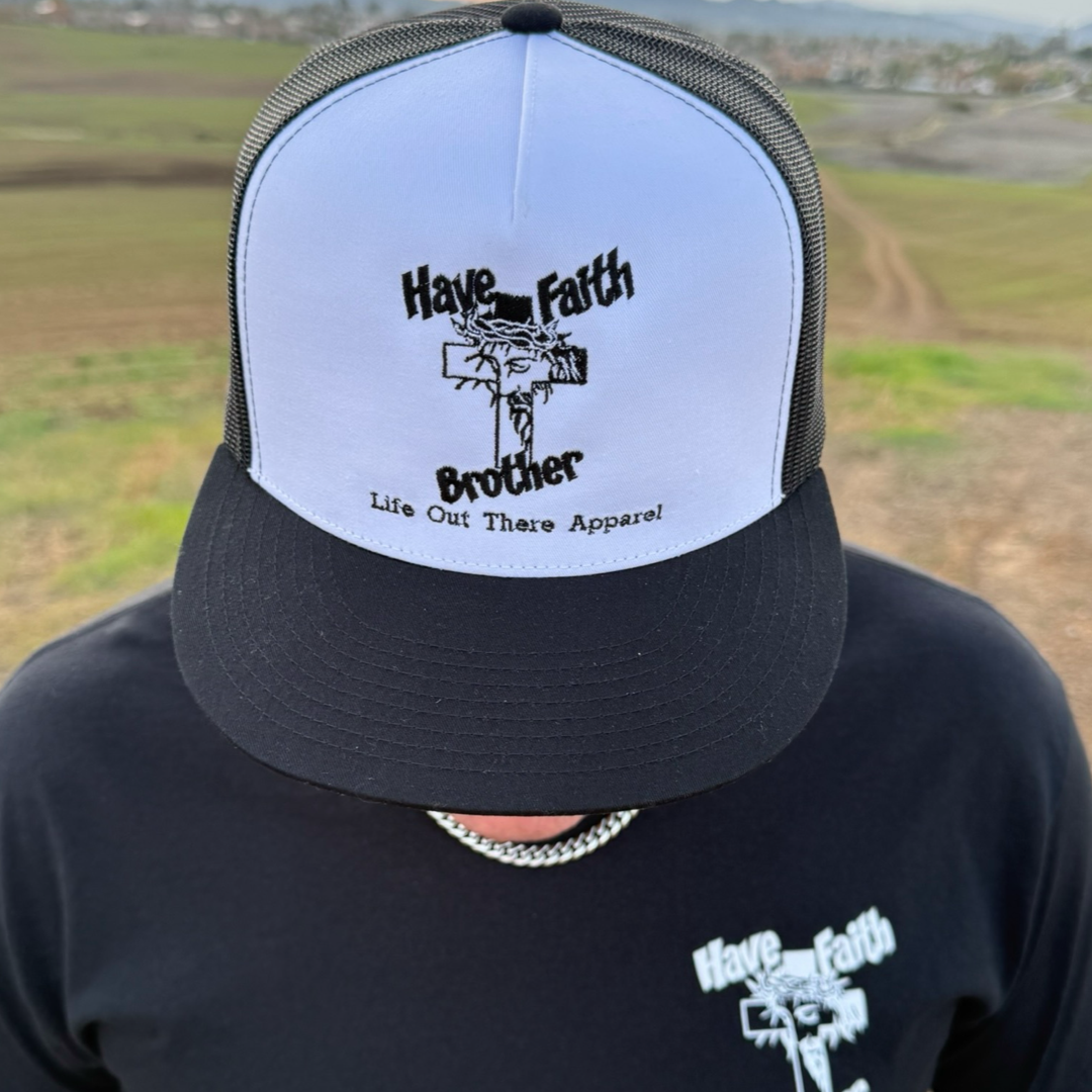 A person stands outdoors on a grassy path wearing the Life Out There Apparel "Have Faith Brother" embroidered flat bill snapback trucker hat (white/black) featuring a cross graphic.