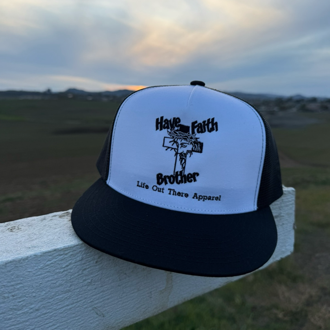 The Have Faith Brother (Embroidered) Flat Bill SnapBack Trucker Hat in White/Black by Life Out There Apparel features a cross and the phrase “Have Faith Brother,” shown resting on a fence with fields and a sunset behind it.