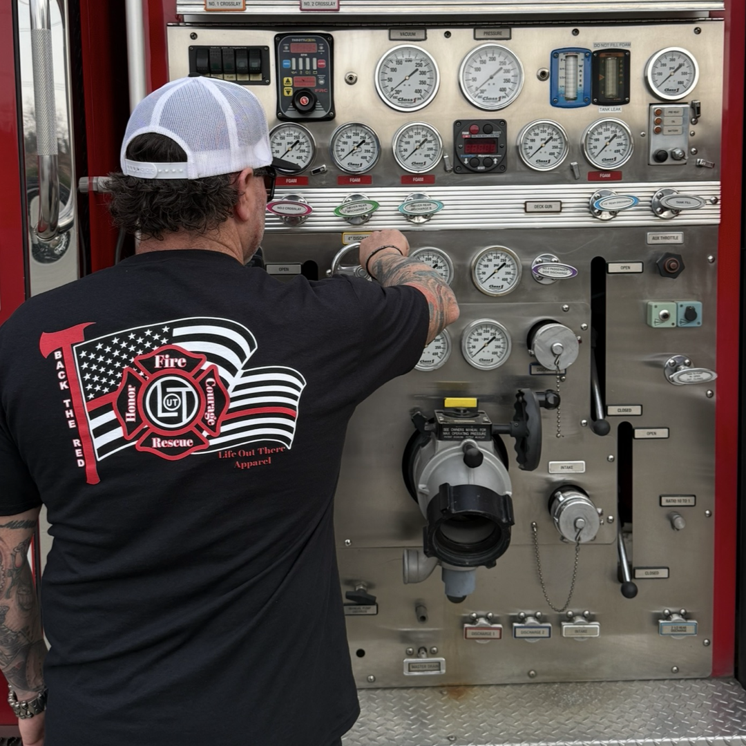 A firefighter in a Life Out There Apparel Back The Red Tee - Black and a white cap skillfully operates the fire truck’s control panel, adjusting gauges and levers on the shiny metal dashboard.