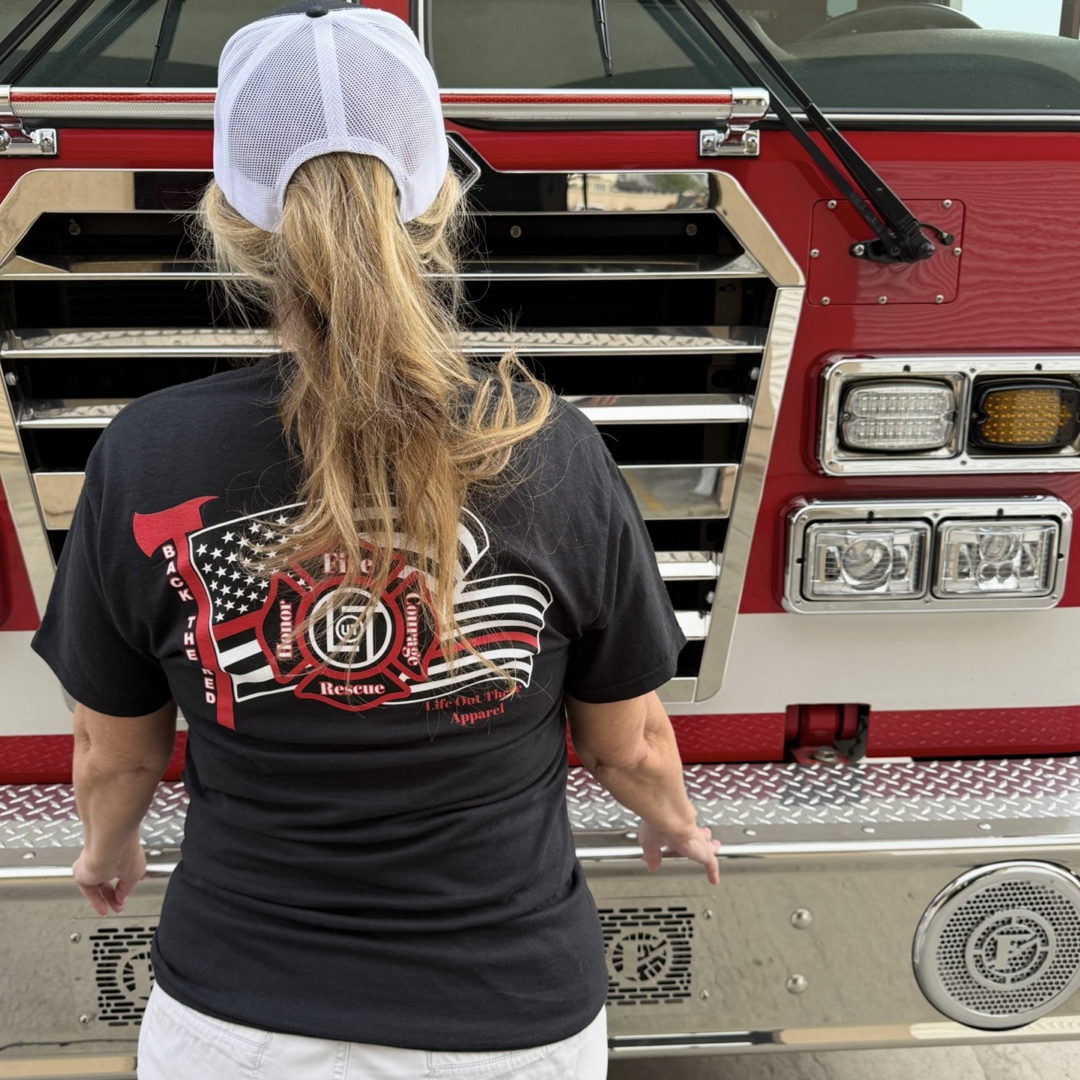A woman in a ponytail and white cap, wearing the Life Out There Apparel Back The Red Tee in black, stands with her back to the camera gripping the chrome bumper of a red fire truck.
