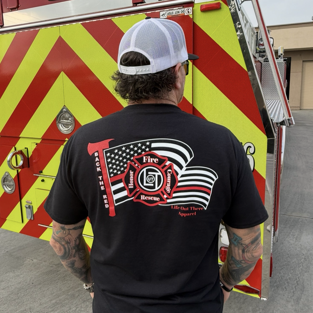 A person in a Life Out There Apparel "Back The Red Tee - Black" featuring an axe, American flag, and “Fire Rescue” and “BACK THE RED” stands before a red and yellow fire truck.
