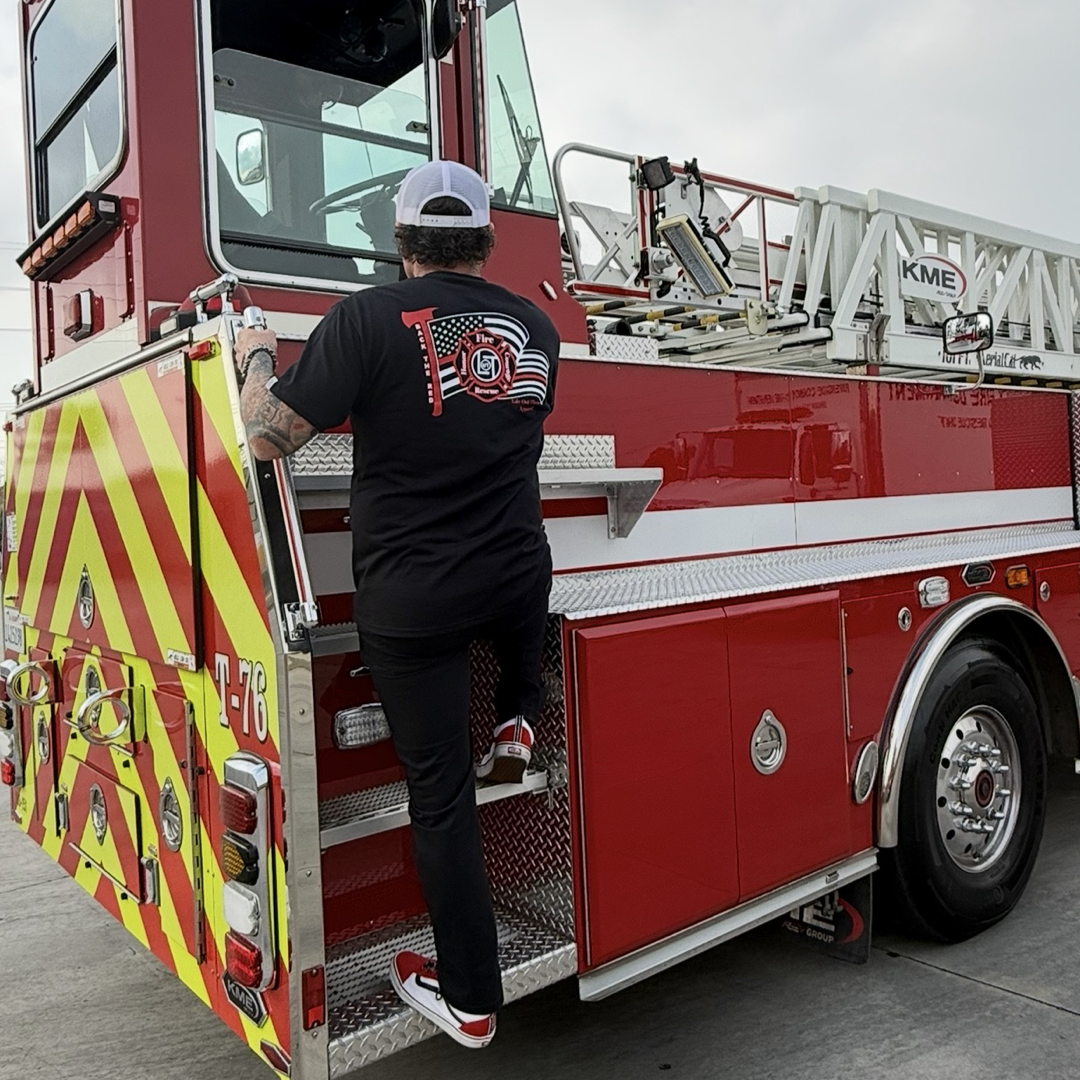 Wearing a Life Out There Apparel Back The Red Tee - Black and cap, someone climbs onto a red fire truck with yellow stripes and American Firefighter pride markings, parked outdoors on pavement.