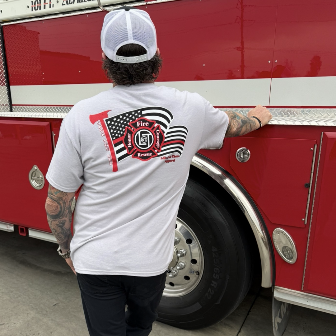 Wearing the Life Out There Apparel Back The Red Tee - Silver, showing a fire rescue logo and U.S. flag, a person stands proudly by a red fire truck—symbolizing pride as an American Firefighter.