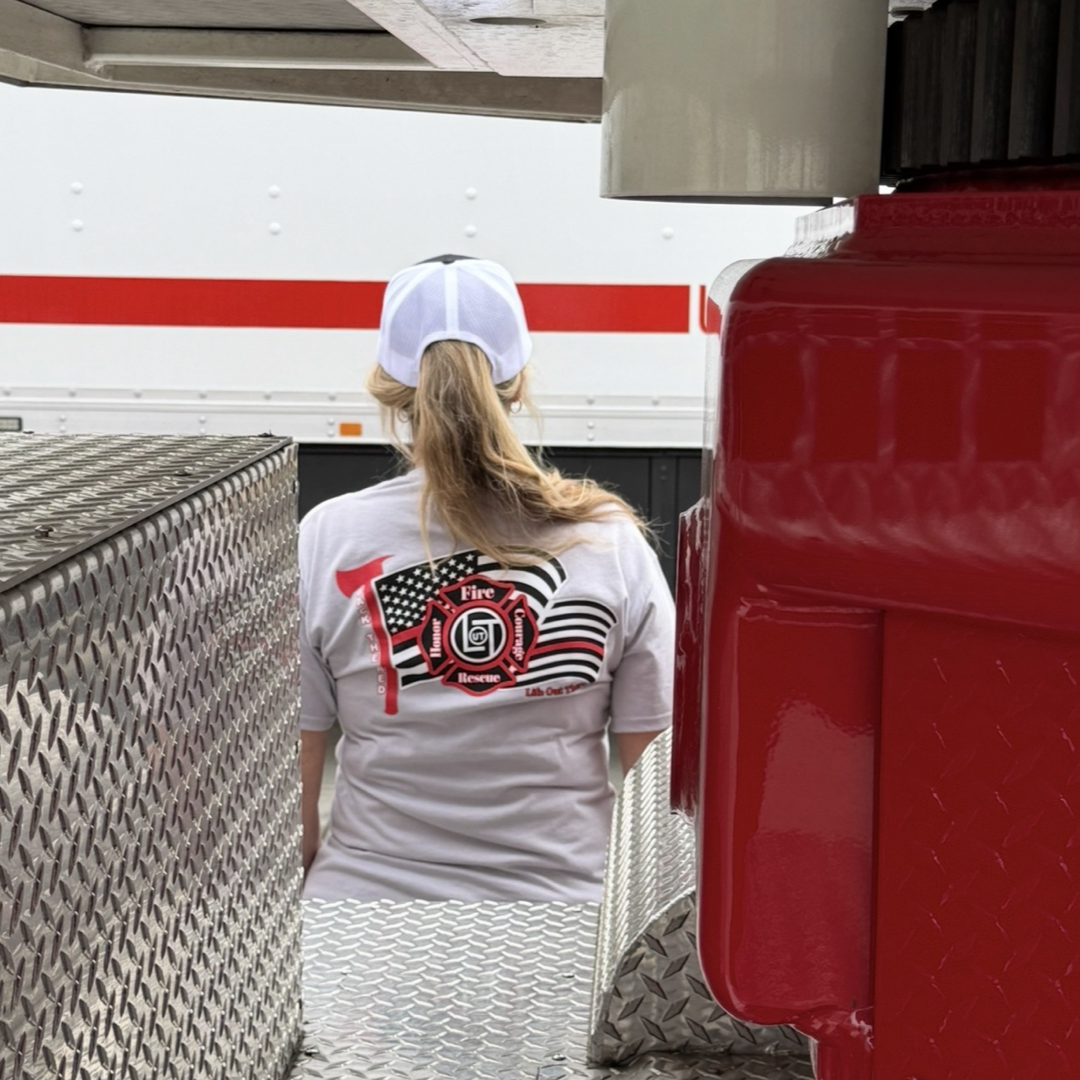 Wearing the Back The Red Tee - Silver from Life Out There Apparel, a person in a white cap stands between shiny metal surfaces near a red fire truck, facing away from the camera to show support for firefighters.