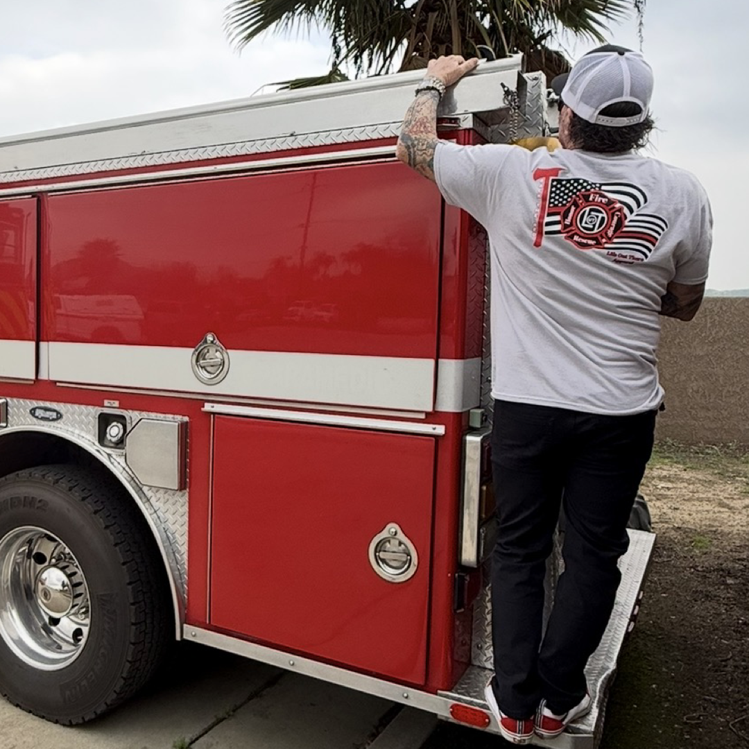 Wearing the Back The Red Tee - Silver by Life Out There Apparel, a firefighter in full gear and white hat climbs a red fire truck, gripping the edge—embodying true American Firefighter spirit.