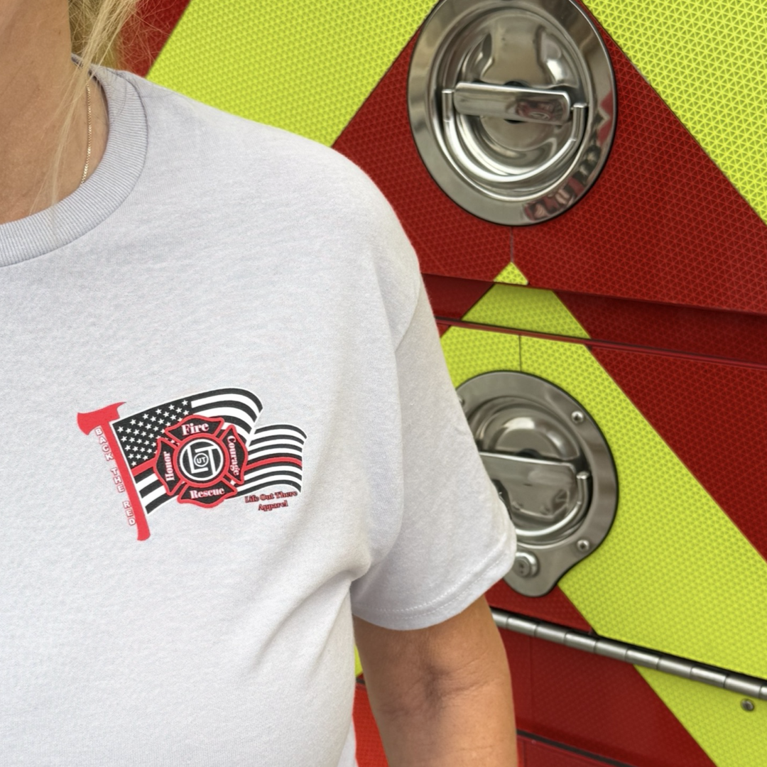 A person in a silver Back The Red Tee by Life Out There Apparel, featuring a firefighter logo and American flag design, stands before a red and yellow fire truck with visible metal handles.