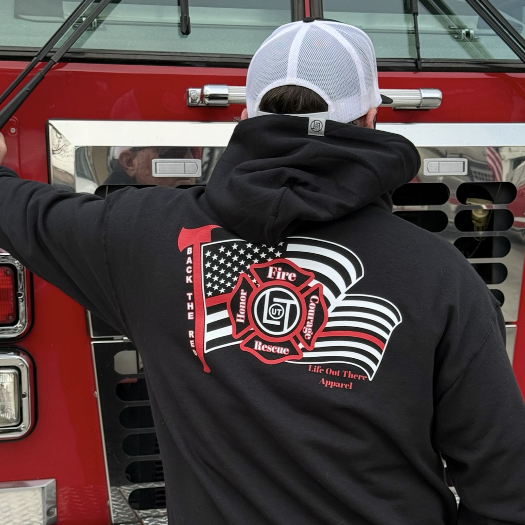 A person in a black Back The Red Hoodie by Life Out There Apparel, featuring a fire rescue emblem and stylized American flag, faces a red fire truck, proudly supporting the Back the Red movement.