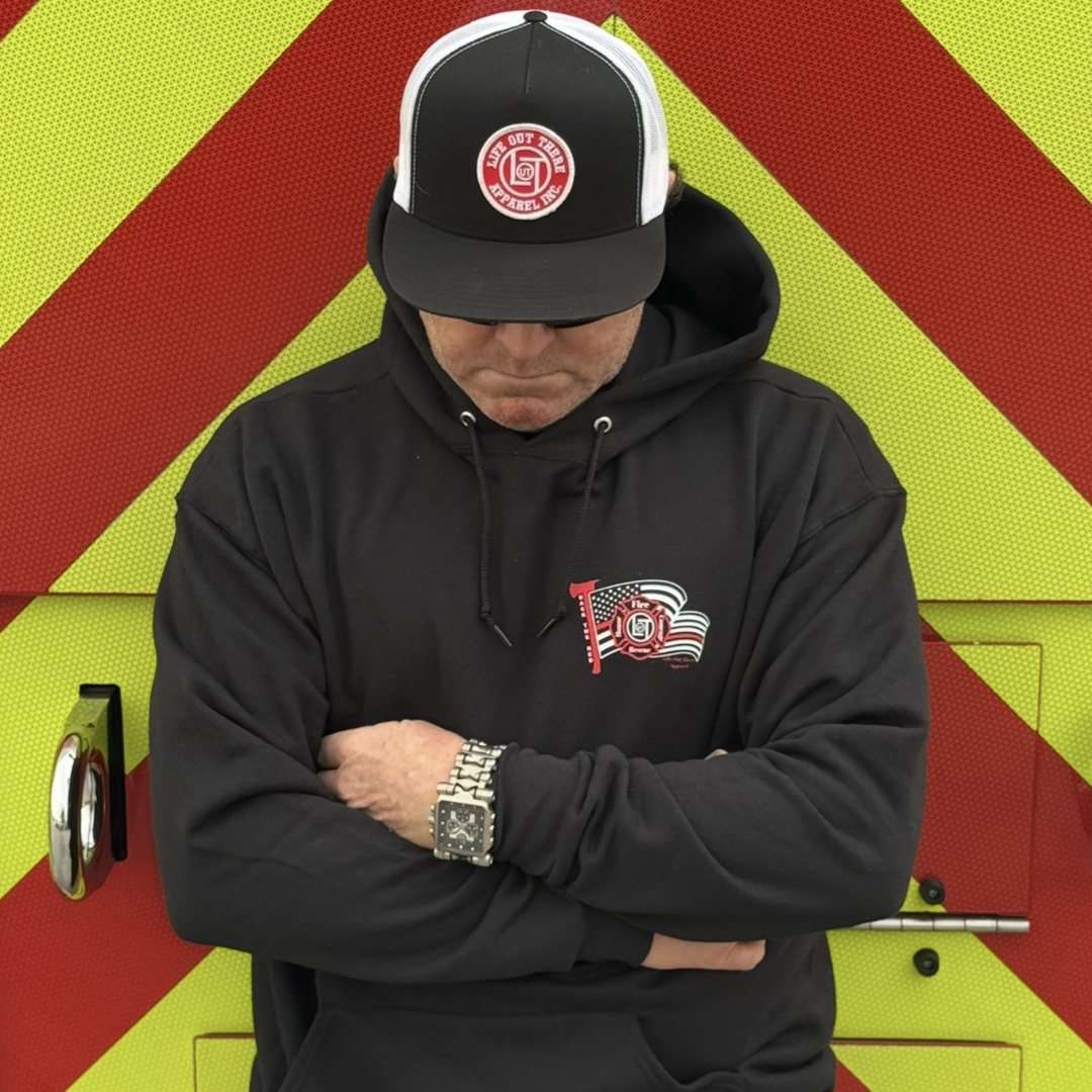 Wearing the Back The Red Hoodie - Black by Life Out There Apparel and a black hat with a fire department logo, a person stands arms crossed in front of a red and yellow chevron-striped vehicle, showcasing firefighter pride.
