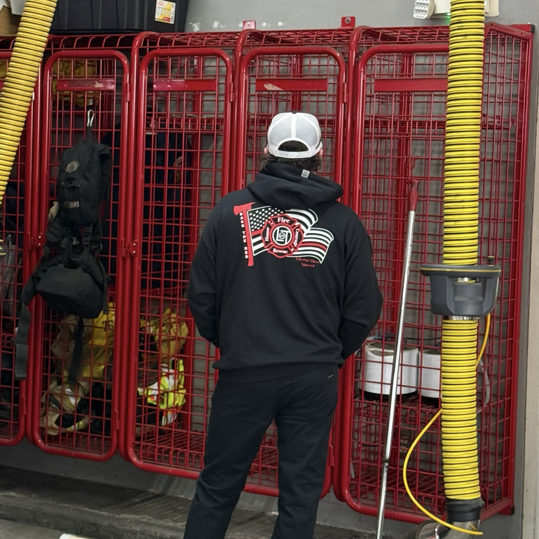 A person wearing the Life Out There Apparel Back The Red Hoodie - Black and a white cap faces red metal lockers in a fire station. Yellow exhaust hoses hang from the ceiling, and firefighter gear is visible inside the lockers.