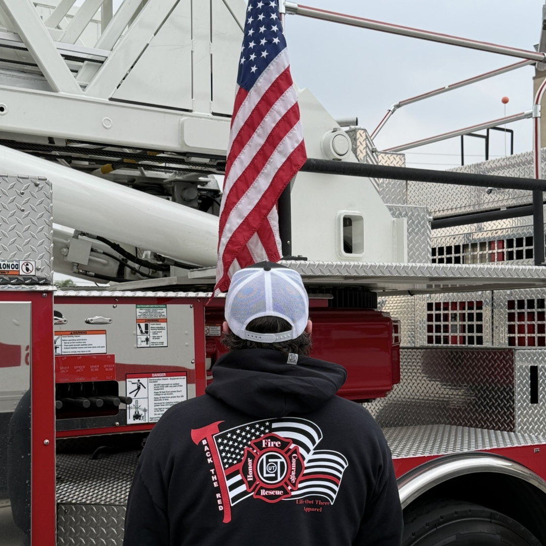 Wearing the Back The Red Hoodie - Black by Life Out There Apparel, a person stands before an American flag on a fire truck, metal gear and ladder behind them—proudly embodying the spirit of the American Firefighter.