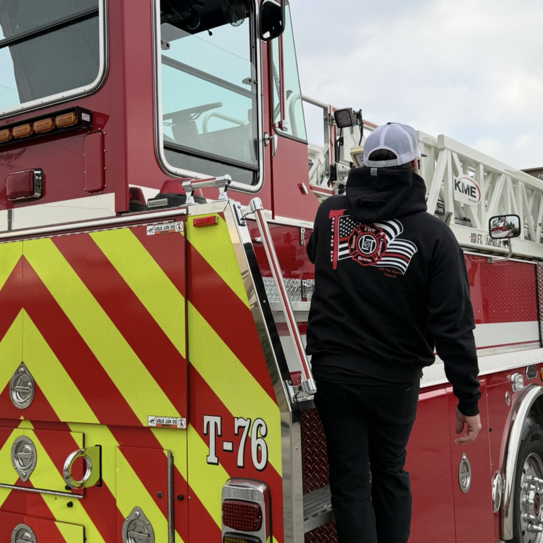Wearing the Life Out There Apparel Back The Red Hoodie - Black, featuring an American flag, a person climbs onto a red fire truck with yellow chevrons and the label T-76 on its side.
