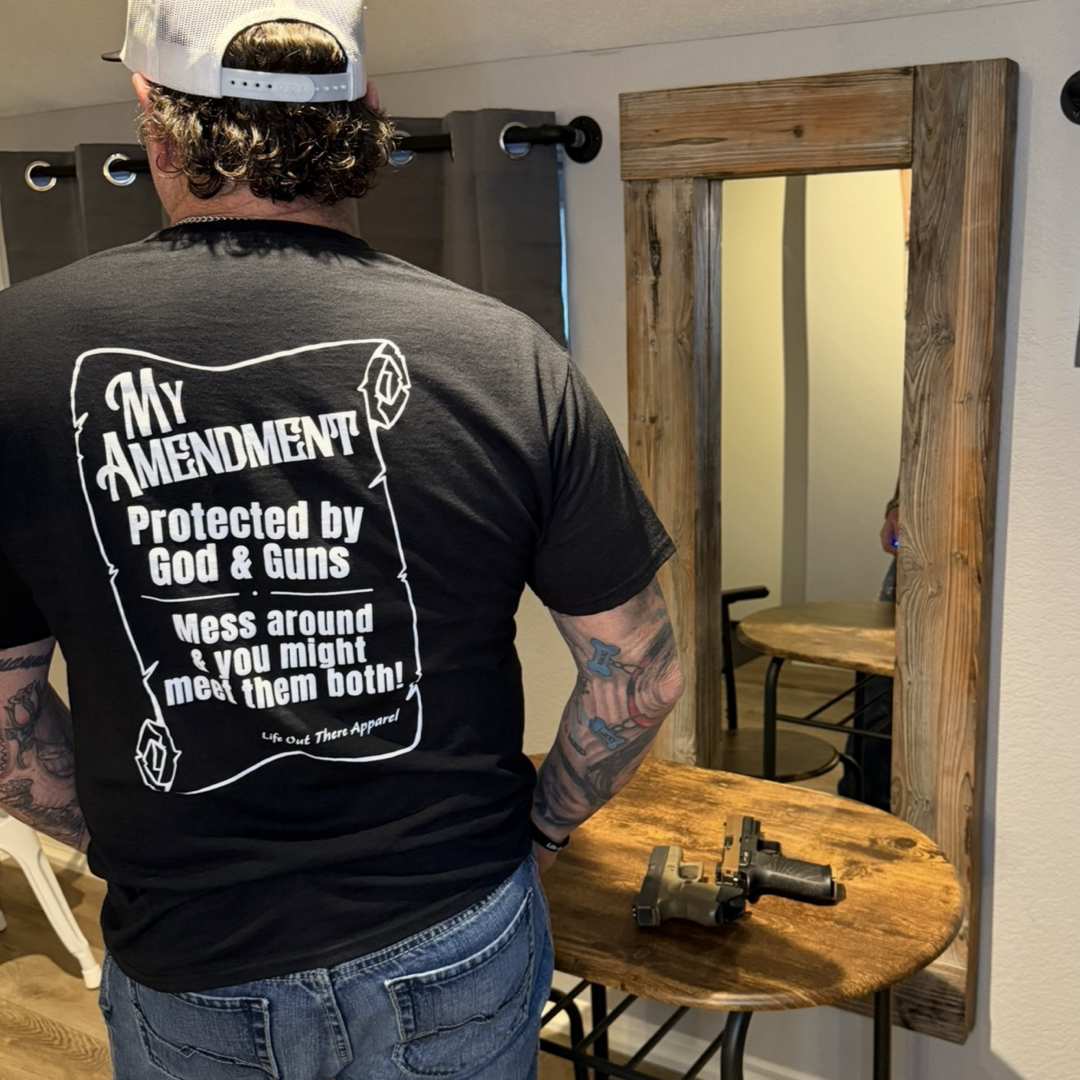A man in a backward cap faces a mirror wearing the "My Amendment Tee - Black" by Life Out There Apparel, featuring the message: “My Amendment Protected by God & Guns. Mess around & you might meet them both!” A handgun sits on a wooden table.