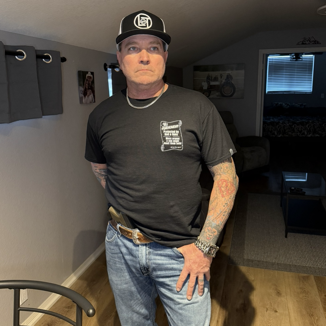 A tattooed man wears the My Amendment Tee - Black by Life Out There Apparel, jeans, a black cap, and a chain necklace. He stands in a room with wooden floors, gray walls, framed pictures, and window light behind him.