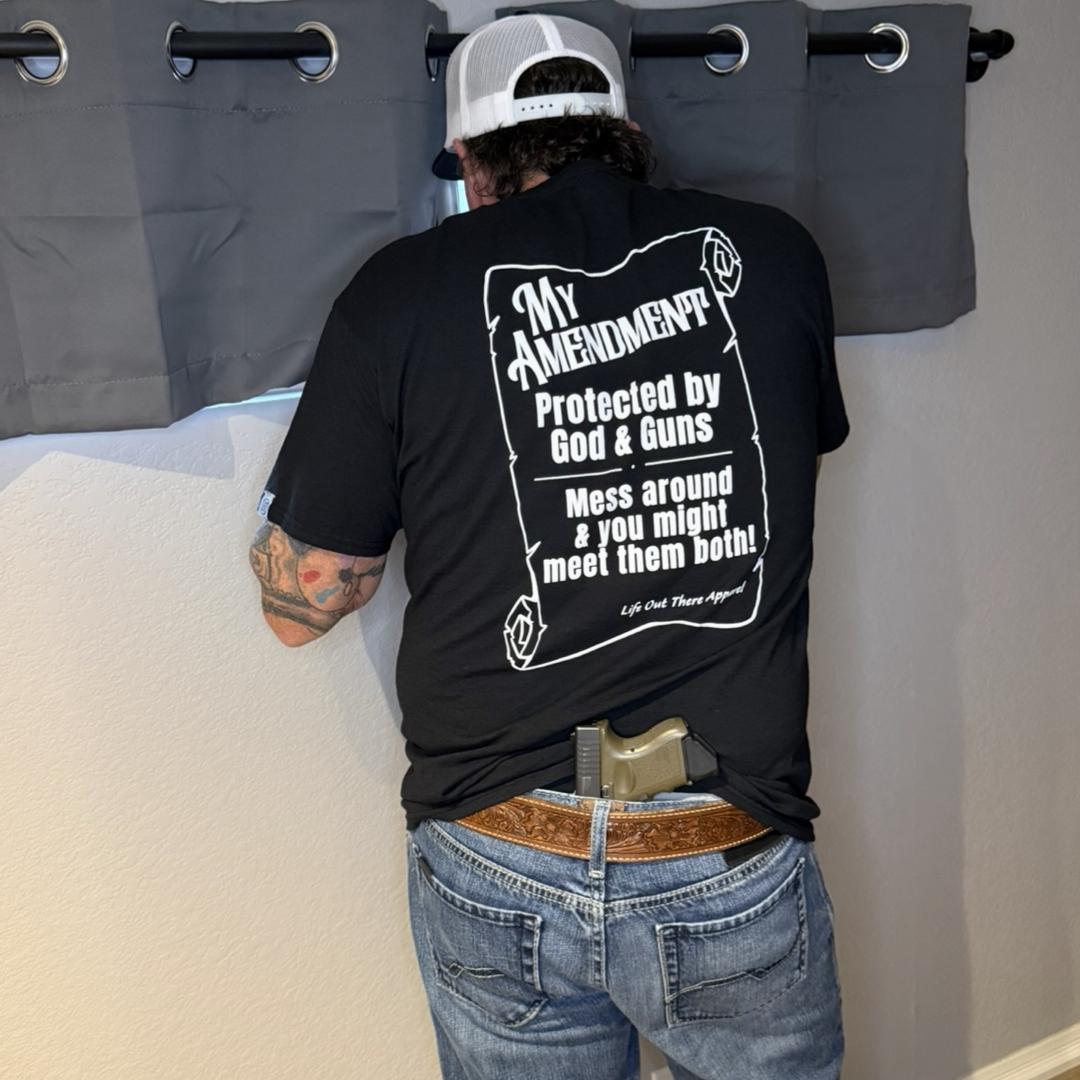 A man in the Life Out There Apparel "My Amendment Tee - Black," jeans, and a white cap looks out a window. A handgun, part of his protection gear, is visible tucked into the back of his waistband.