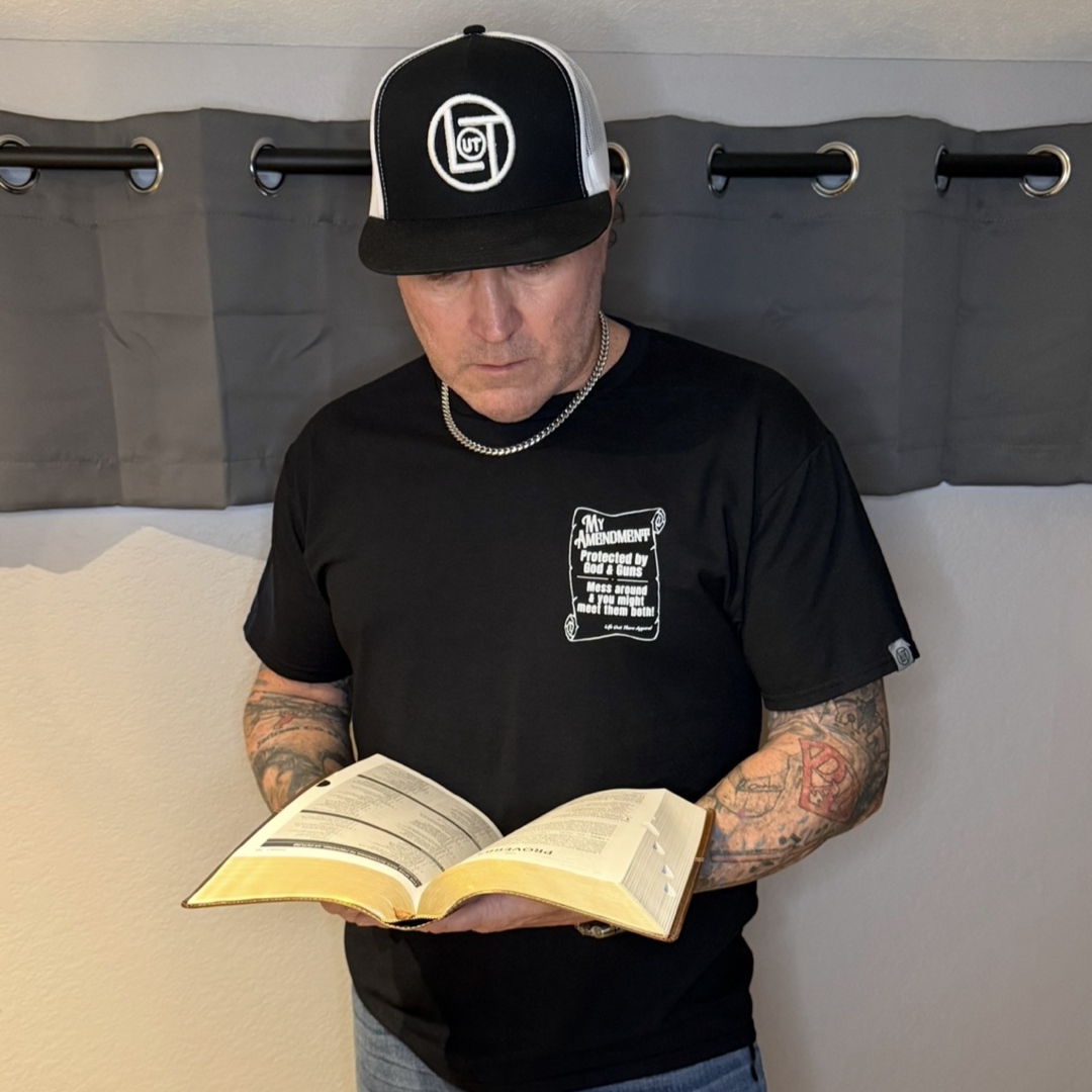 A man in the My Amendment Tee - Black by Life Out There Apparel stands indoors with tattoos on his arms, holding and reading an open book. He faces downward, and a gray curtain is behind him.