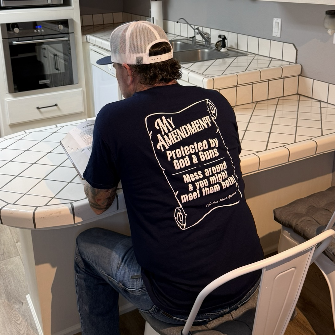 A man in a Life Out There Apparel My Amendment Tee - Navy, featuring the bold statement "Protected by God & Guns—mess around & you might meet them both," sits at a tiled kitchen counter reading a document, wearing a white cap.