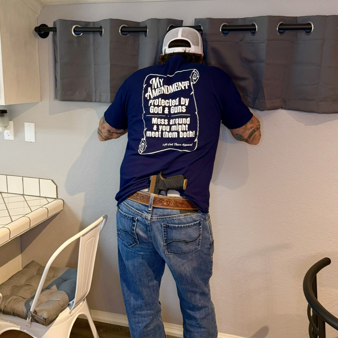 A man in jeans and a navy My Amendment Tee by Life Out There Apparel stands facing a window, adjusting gray curtains with a handgun tucked in his waistband. Protector apparel from this faith-based brand adds purpose to his stance. A white chair sits nearby.