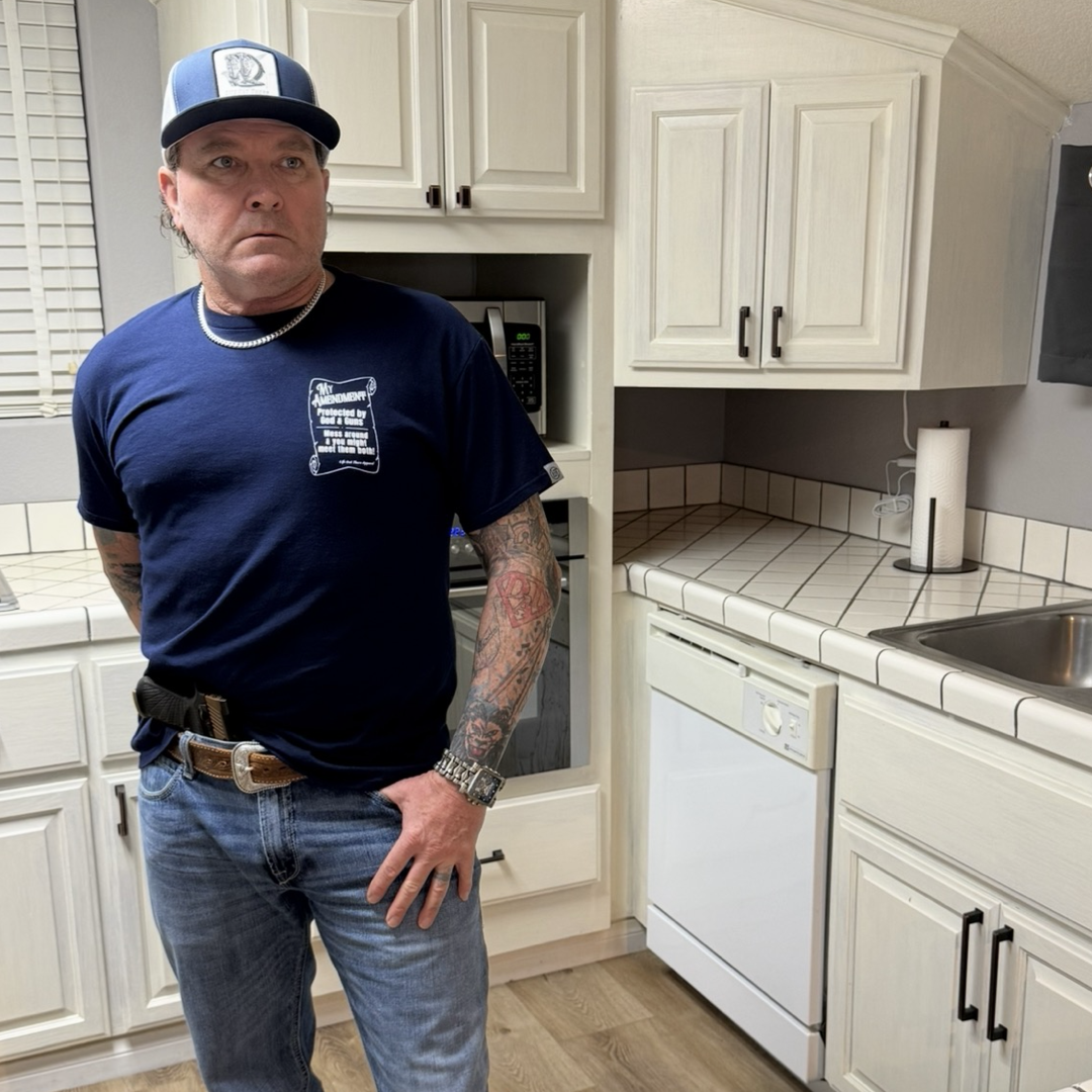 A man with tattooed arms, in jeans, a watch, a blue-and-white cap, and the My Amendment Tee - Navy from Life Out There Apparel stands in a white-tiled kitchen with cream cabinets, a dishwasher, and a microwave.