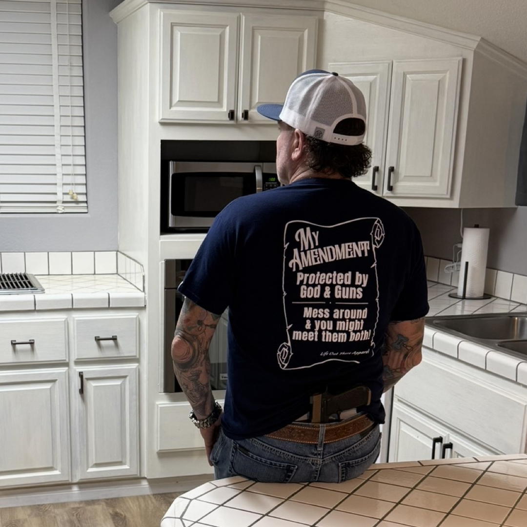 A man stands in a white kitchen with his back to the camera, wearing the navy My Amendment Tee from Life Out There Apparel—a faith-based shirt featuring God & Guns and Second Amendment text, warning others not to mess around.