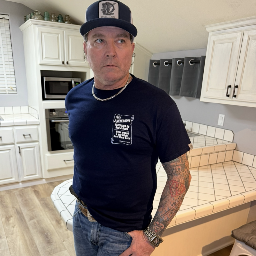 A tattooed man in a navy My Amendment Tee by Life Out There Apparel and a cap stands in a white kitchen with tiled counters and wooden floors, resting one hand on his hip as he looks to his left.