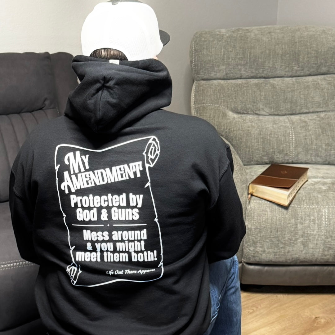 Wearing a black My Amendment Hoodie by Life Out There Apparel, a person in a white cap sits on a couch. The hoodie’s back reads: “Protected by God & Guns. Mess around & you might meet them both!” A closed Bible lies on the table.
