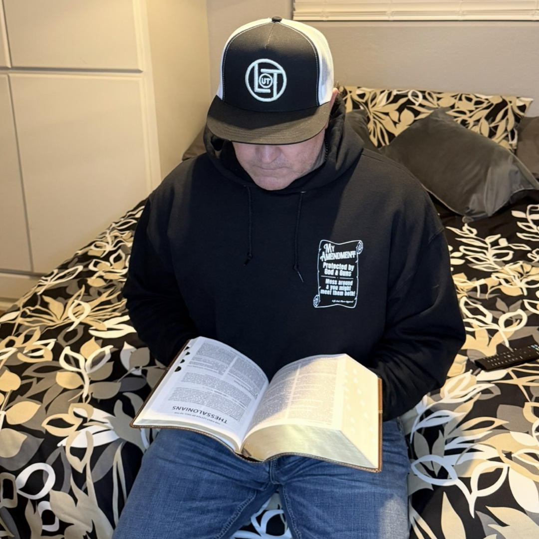 Wearing the My Amendment Hoodie - Black by Life Out There Apparel and a cap, a person sits on a neatly made, well-lit bed with patterned bedding, reading a book. A TV remote rests nearby.