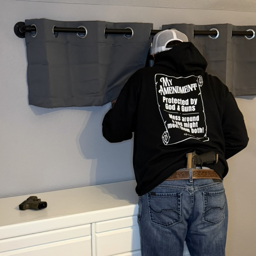 Wearing the Life Out There Apparel My Amendment Hoodie - Black, a person hangs gray curtains with a pistol holstered at their waist and another placed on a white dresser.
