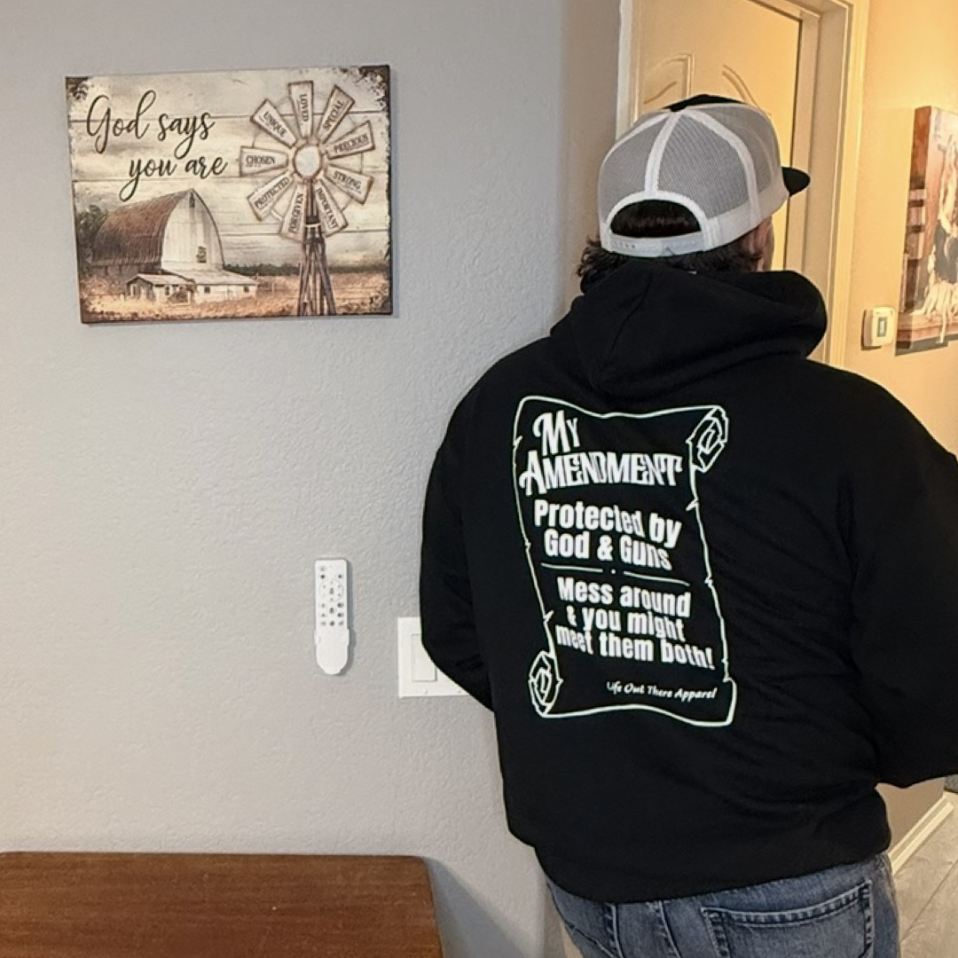 Wearing the My Amendment Hoodie - Black by Life Out There Apparel, a person stands indoors facing a wall with a farmhouse-style "God says you are" sign and a remote attached beneath it.