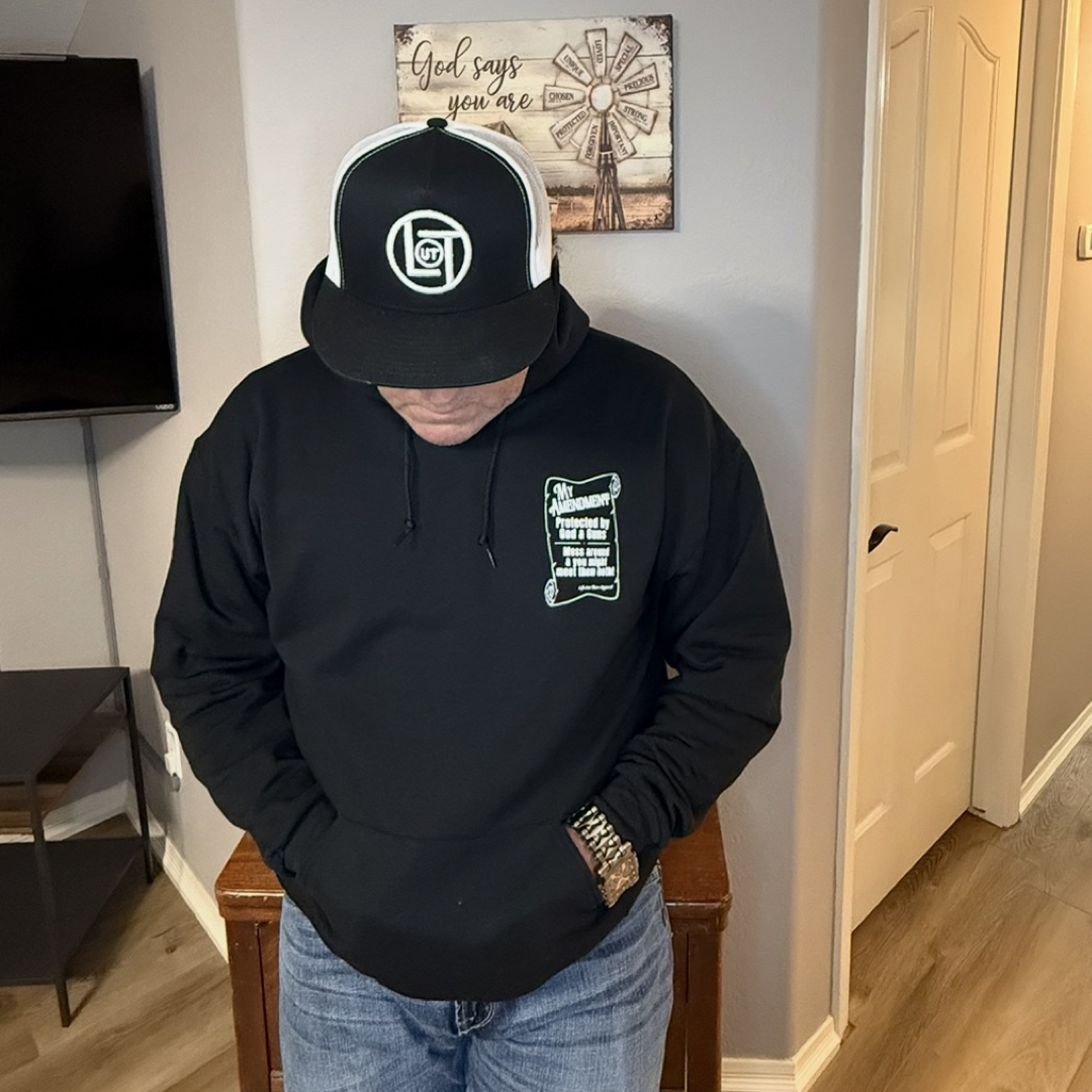 A person wearing Life Out There Apparel’s My Amendment Hoodie in black stands indoors, head down with hands in pockets. Above them is a wall sign reading "God says you are" beside a windmill picture.
