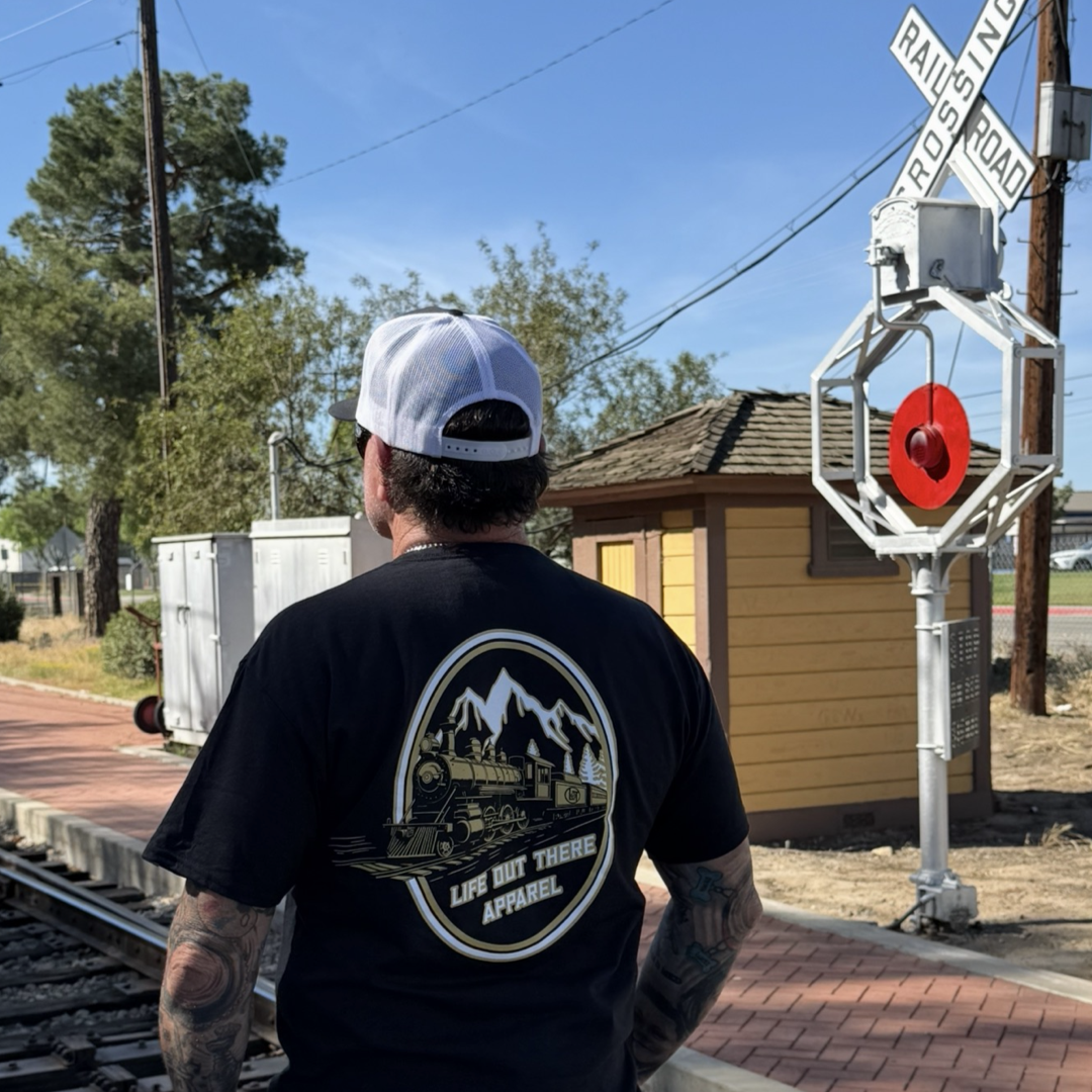 A man in a black Don’t Get Derailed Tee by Life Out There Apparel and a white cap stands near train tracks by a railroad crossing sign, soaking up the sun beside a small yellow building—a reminder: Don’t Get Derailed.