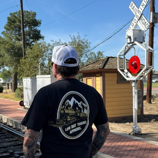 A man in a black Don’t Get Derailed Tee by Life Out There Apparel and a white cap stands near train tracks by a railroad crossing sign, soaking up the sun beside a small yellow building—a reminder: Don’t Get Derailed.