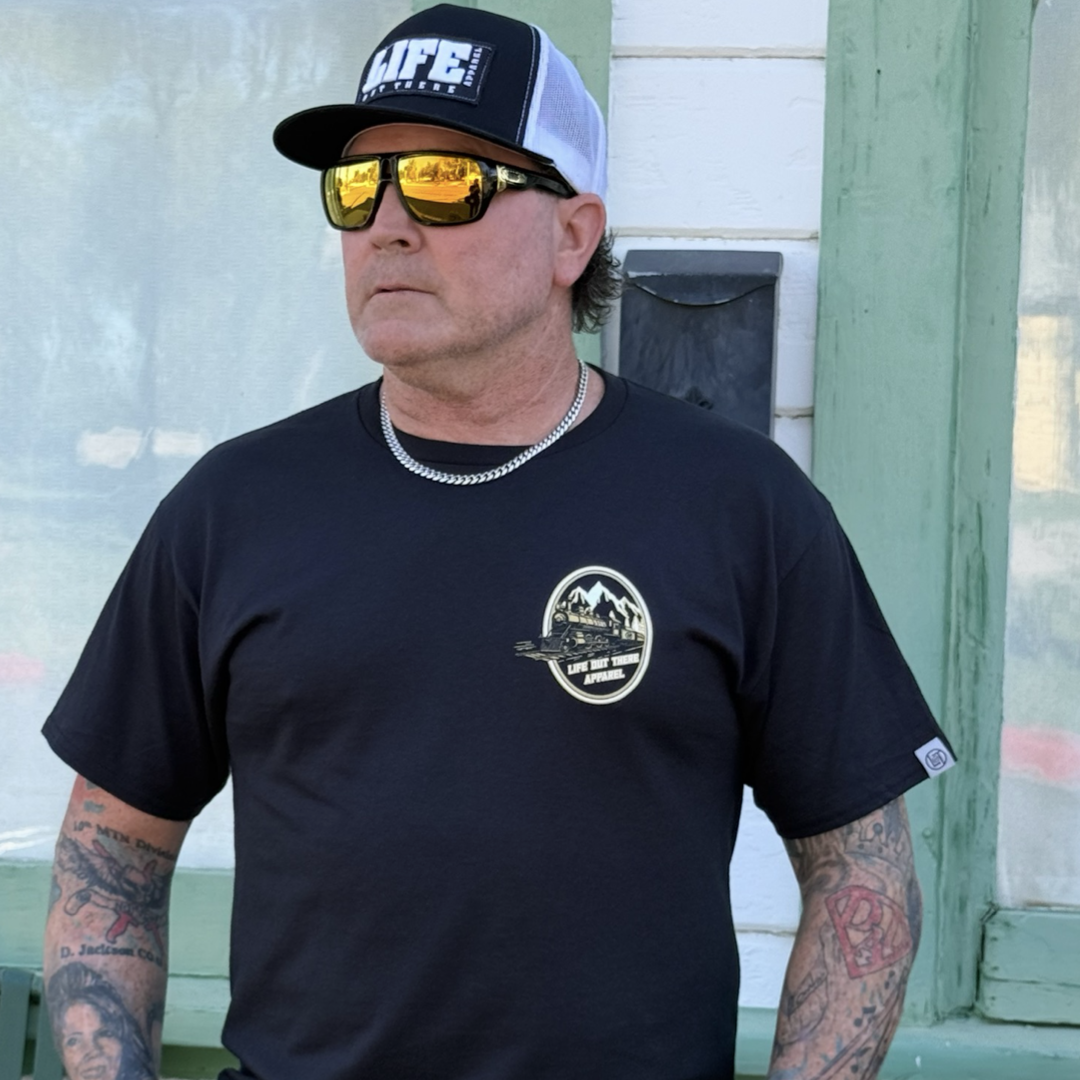 A tattooed man wearing the Don’t Get Derailed Tee in black and a black and white LIFE trucker hat, both by Life Out There Apparel, plus a chain necklace and mirrored sunglasses, stands looking left in front of a green-trimmed building.
