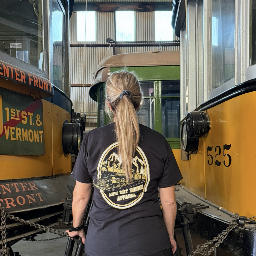A woman with long blonde hair in a ponytail wears the black Don’t Get Derailed Tee by Life Out There Apparel, which features trains and the phrase "Don't Get Derailed," as she stands between two vintage streetcars in a train museum.