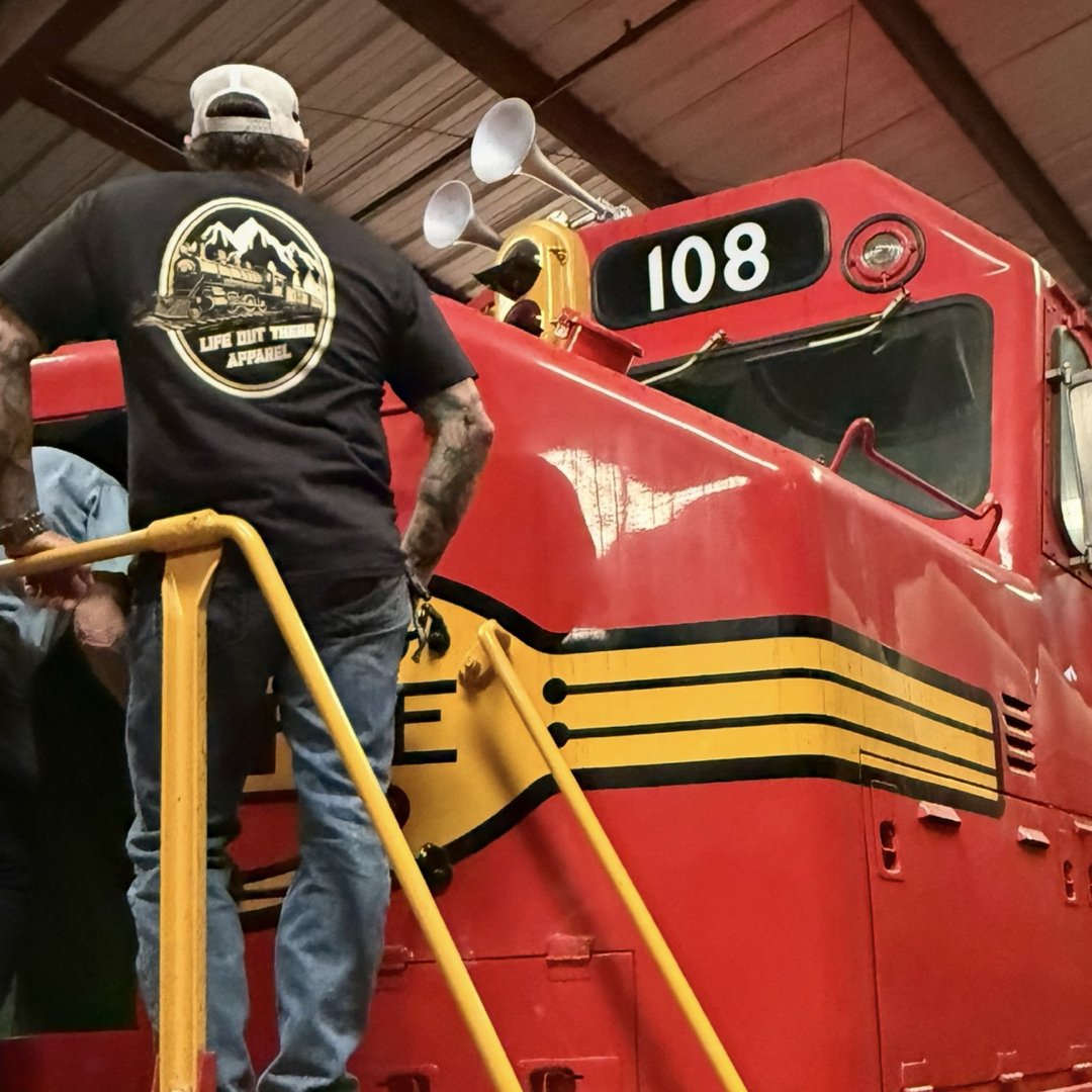 Wearing the Life Out There Apparel Don’t Get Derailed Tee - Black, a man stands on yellow stairs by bright red locomotive 108 inside a railway building, capturing the spirit of Locomotive Engineers—Don’t Get Derailed.