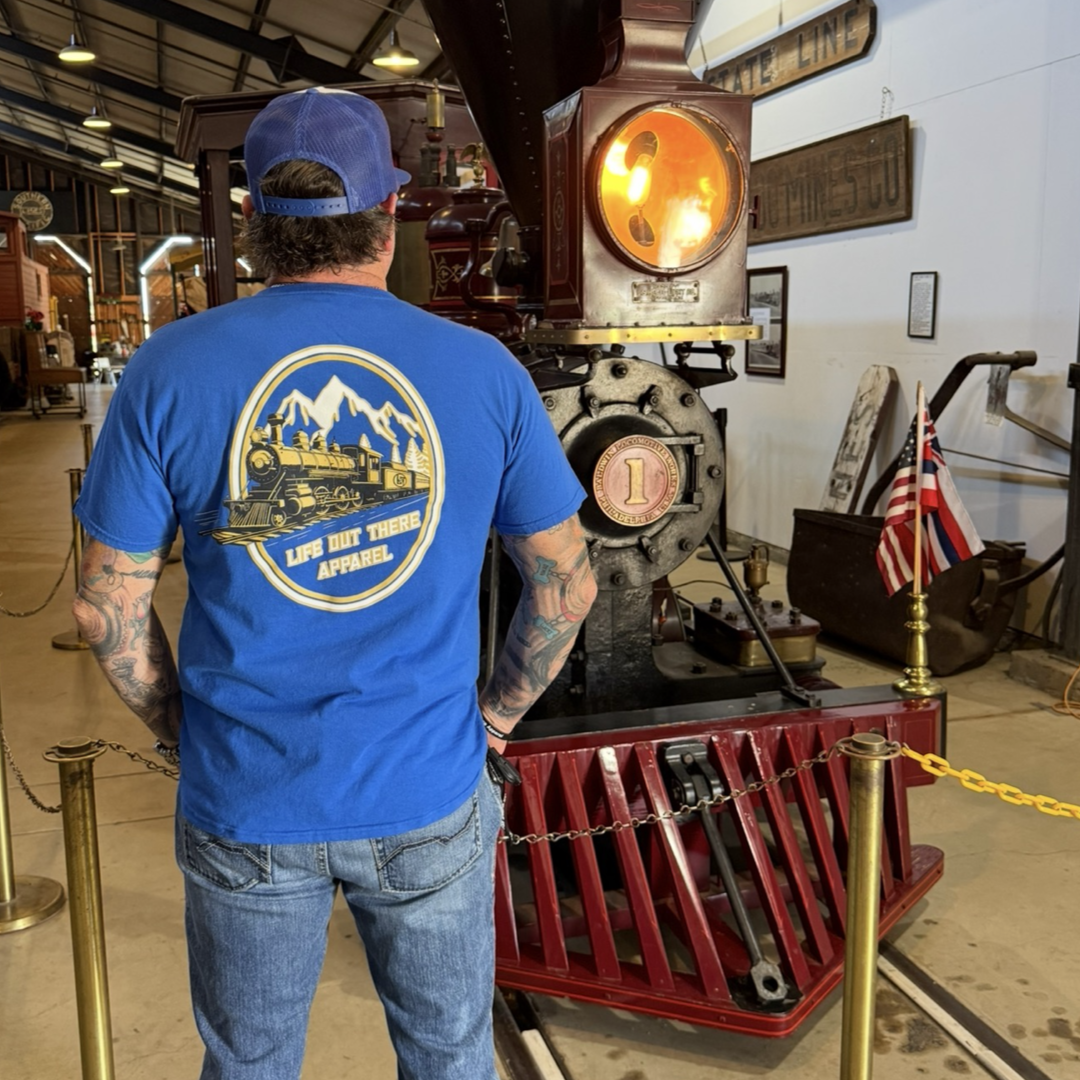 A man in a royal blue Don’t Get Derailed Tee by Life Out There Apparel stands facing a vintage steam locomotive inside a museum, with an American flag and historical signs in the background.