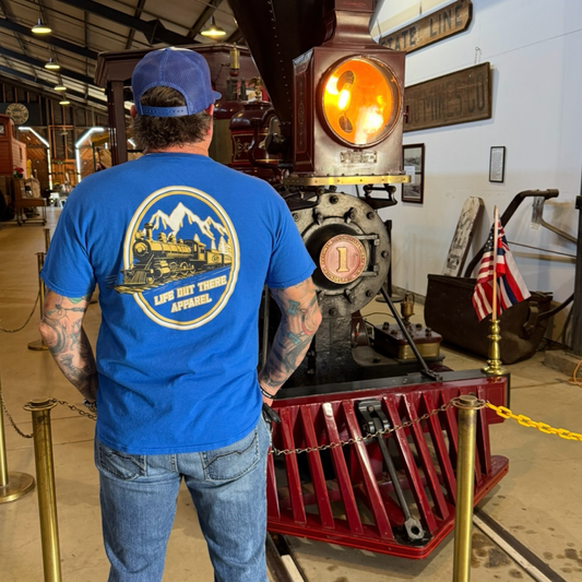 A man in a royal blue Don’t Get Derailed Tee by Life Out There Apparel stands facing a vintage steam locomotive inside a museum, with an American flag and historical signs in the background.