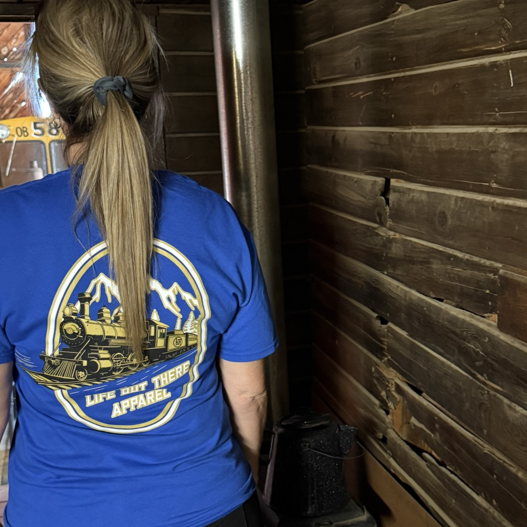 A person with light brown hair in a ponytail wears the royal blue "Don’t Get Derailed Tee" by Life Out There Apparel, featuring a train and mountain graphic, while standing in a rustic wooden cabin near a metal stove—ideal for locomotive fans.