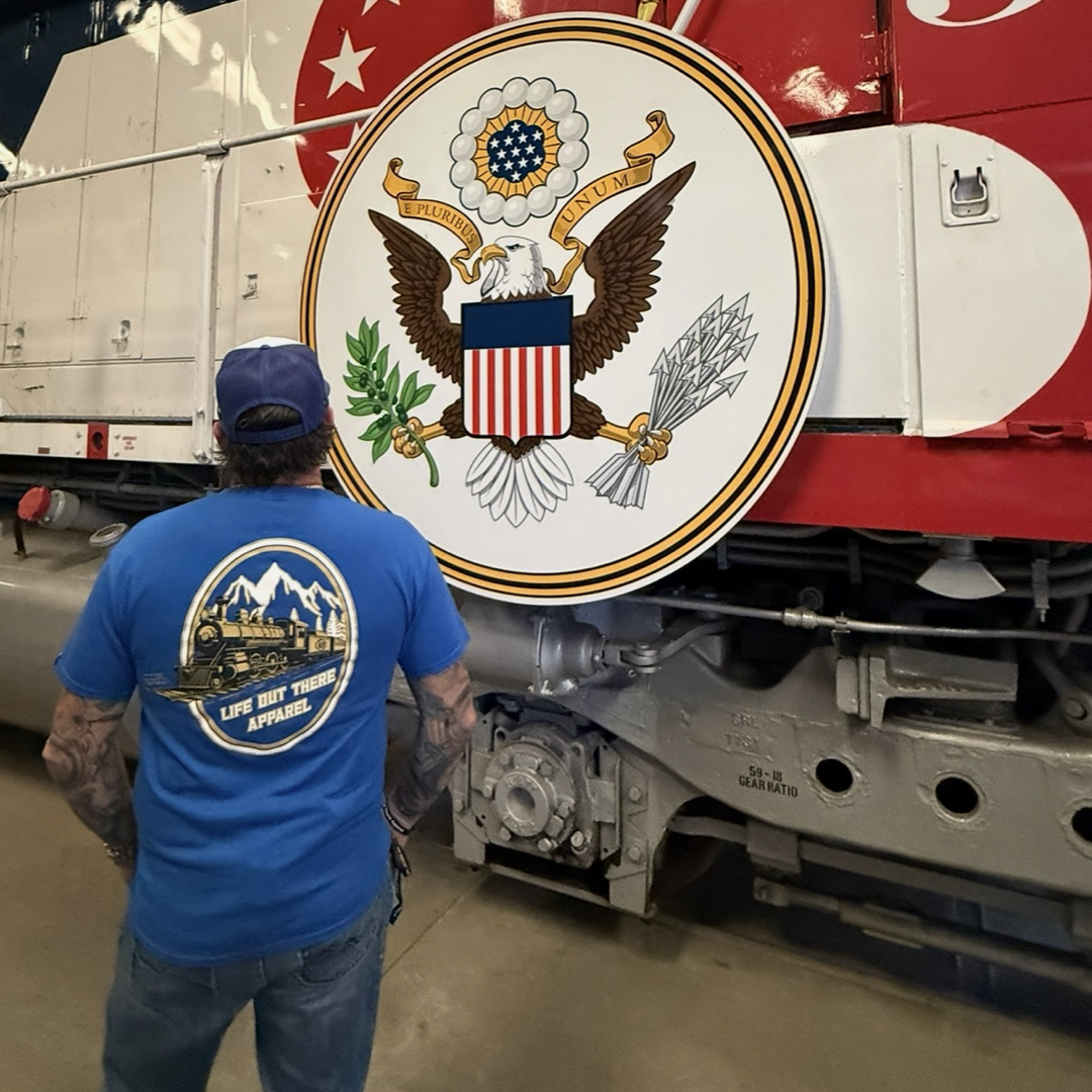 A man in a royal blue "Don’t Get Derailed Tee" by Life Out There Apparel stands before a large train engine displaying the Great Seal of the United States, honoring Locomotive Engineers.