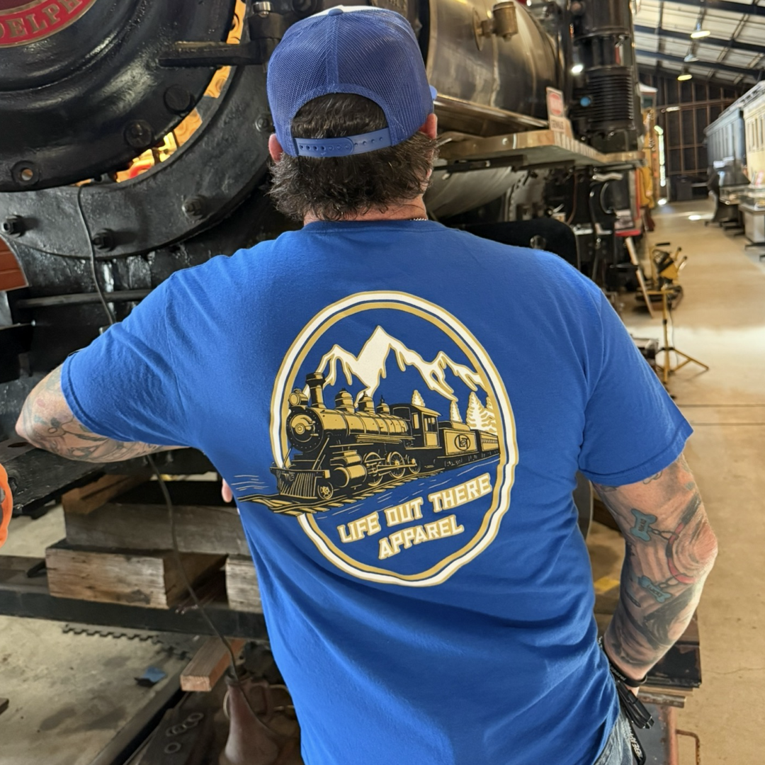 A person in a blue hat and Life Out There Apparel’s Unisex Don’t Get Derailed Tee in Royal Blue stands before a vintage steam locomotive at a train museum. This tee, made for Locomotive Engineers, features a train-and-mountain graphic.