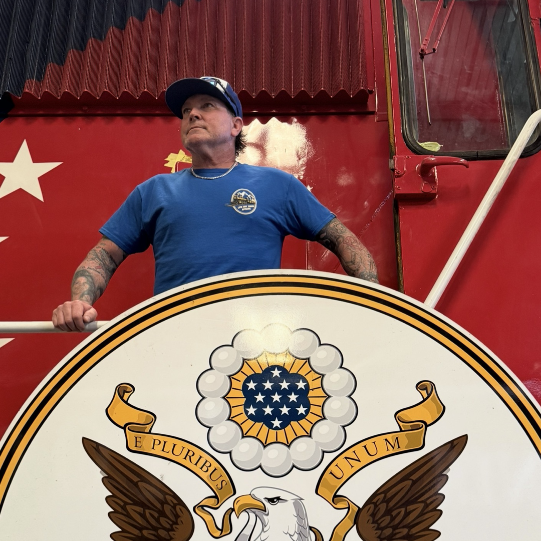 A man in a blue shirt and cap stands behind a large U.S. seal emblem, hands on the railing. The patriotic red wall with white stars is the ideal backdrop for the “Don’t Get Derailed Tee - Royal Blue” by Life Out There Apparel.