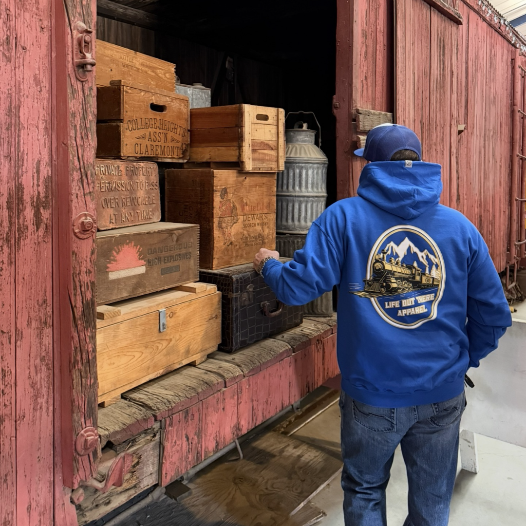 Wearing the Don’t Get Derailed Hoodie in royal blue by Life Out There Apparel, someone stands by a vintage red train car, eyeing classic wooden crates and metal containers—ideal for locomotive enthusiasts and fans of motivational gear.