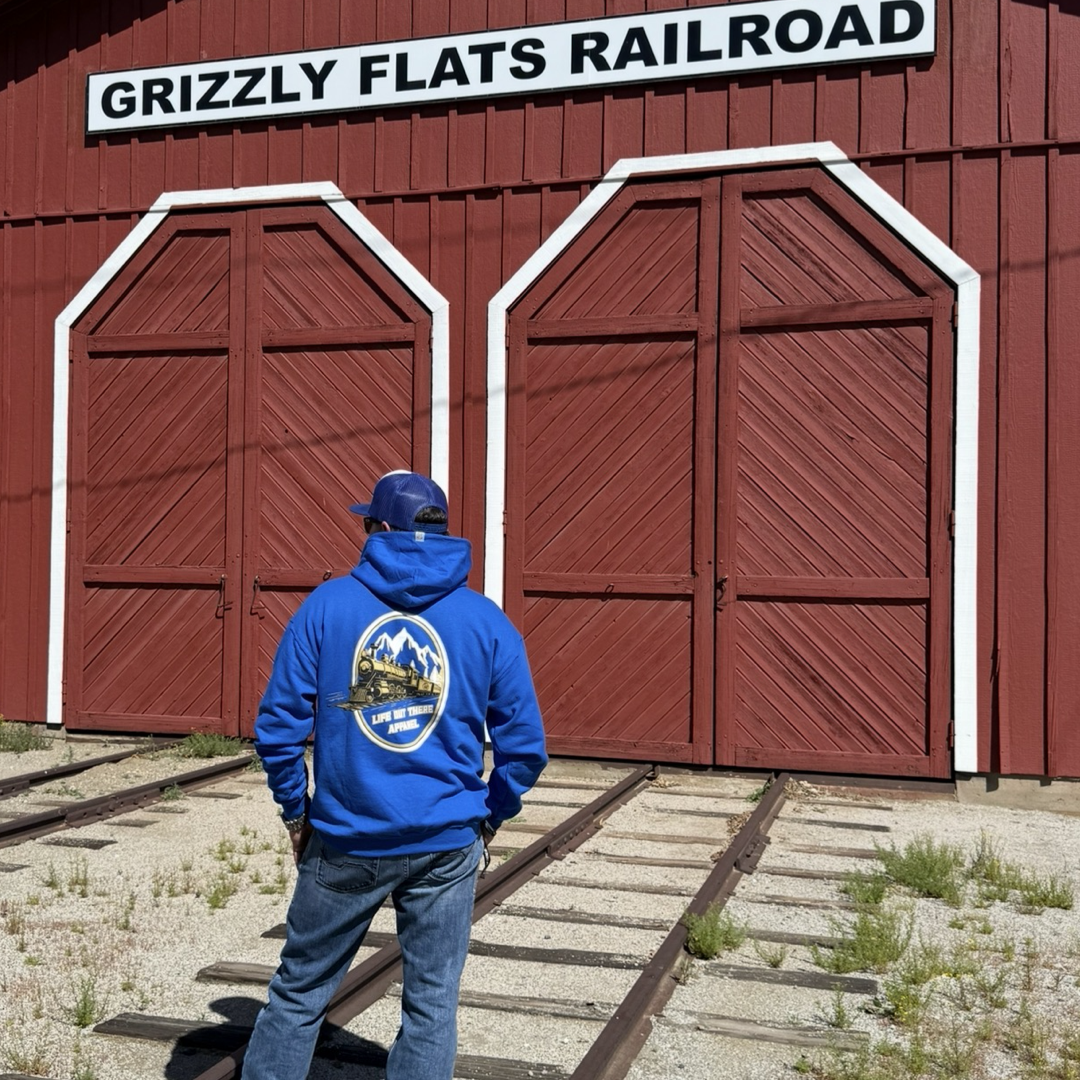 Wearing the “Don’t Get Derailed Hoodie - Royal Blue” by Life Out There Apparel, a person in jeans stands on railroad tracks in front of red barn doors beneath the Grizzly Flats Railroad sign.