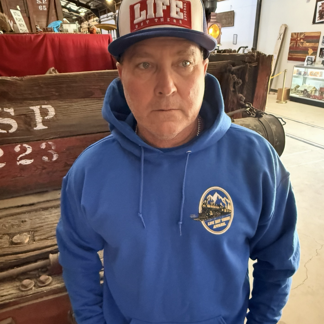 A man in a Don’t Get Derailed Hoodie - Royal Blue by Life Out There Apparel and a LIFE Out Here cap stands indoors near vintage wooden equipment, with motivational style and retro decor in the background.