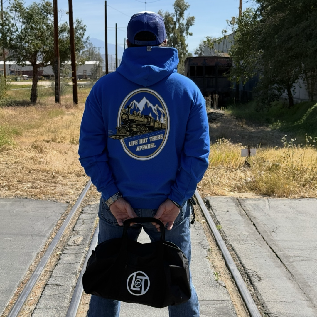 A person in a royal blue Don’t Get Derailed Hoodie by Life Out There Apparel stands on railroad tracks, holding a black duffel bag behind their back. The hoodie features a mountain and train graphic.