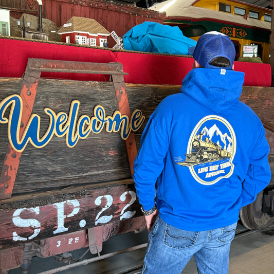 Wearing the Don’t Get Derailed Hoodie in Royal Blue by Life Out There Apparel, a person stands beside vintage train car S.P. 22 and a Welcome sign inside a museum dedicated to Locomotive Engineers.