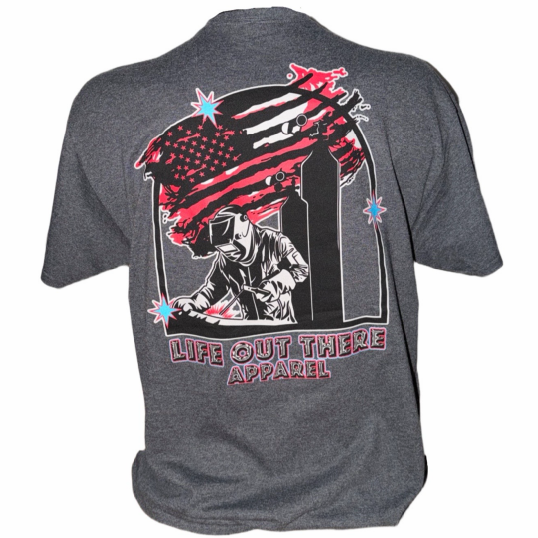 The Tack It Tee - Black Heather by Life Out There Apparel is a unisex shirt featuring a welder graphic, a red and black stylized American flag with stars, and "Life Out There Apparel" plus "TACK IT" text on the back.