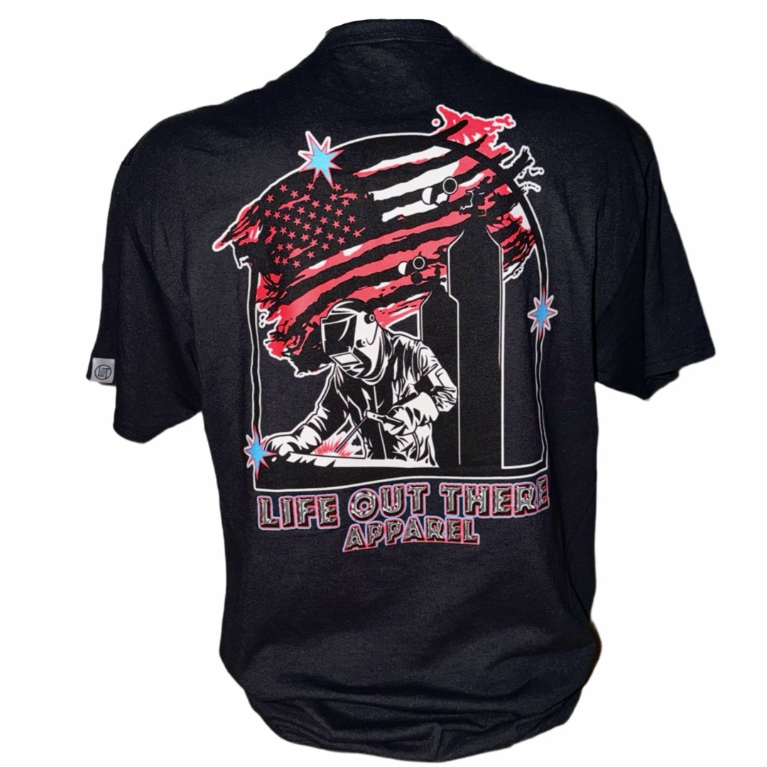 The Tack It Tee - Black by Life Out There Apparel features a graphic of a hoodie-clad figure spray painting with an American flag and city skyline—ideal for welders. Proudly show your style and TACK IT on!.