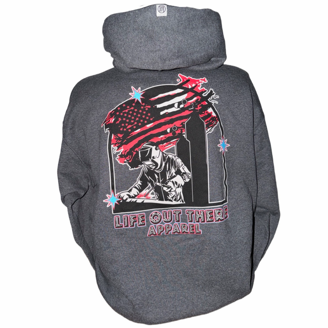 The Tack It Hoodie - Black Heather by Life Out There Apparel is a unisex hoodie featuring a firefighter graphic, twin black towers, distressed flag, and bold LIFE OUT THERE APPAREL text—ideal for welders or anyone who loves to TACK IT.