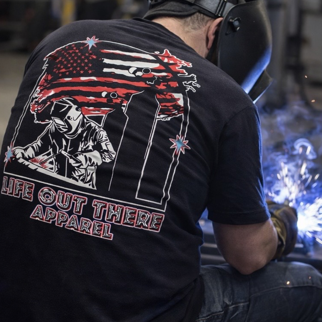 A person in a black Tack It Tee by Life Out There Apparel, featuring an American flag and welder graphic, is welding with bright sparks flying.
