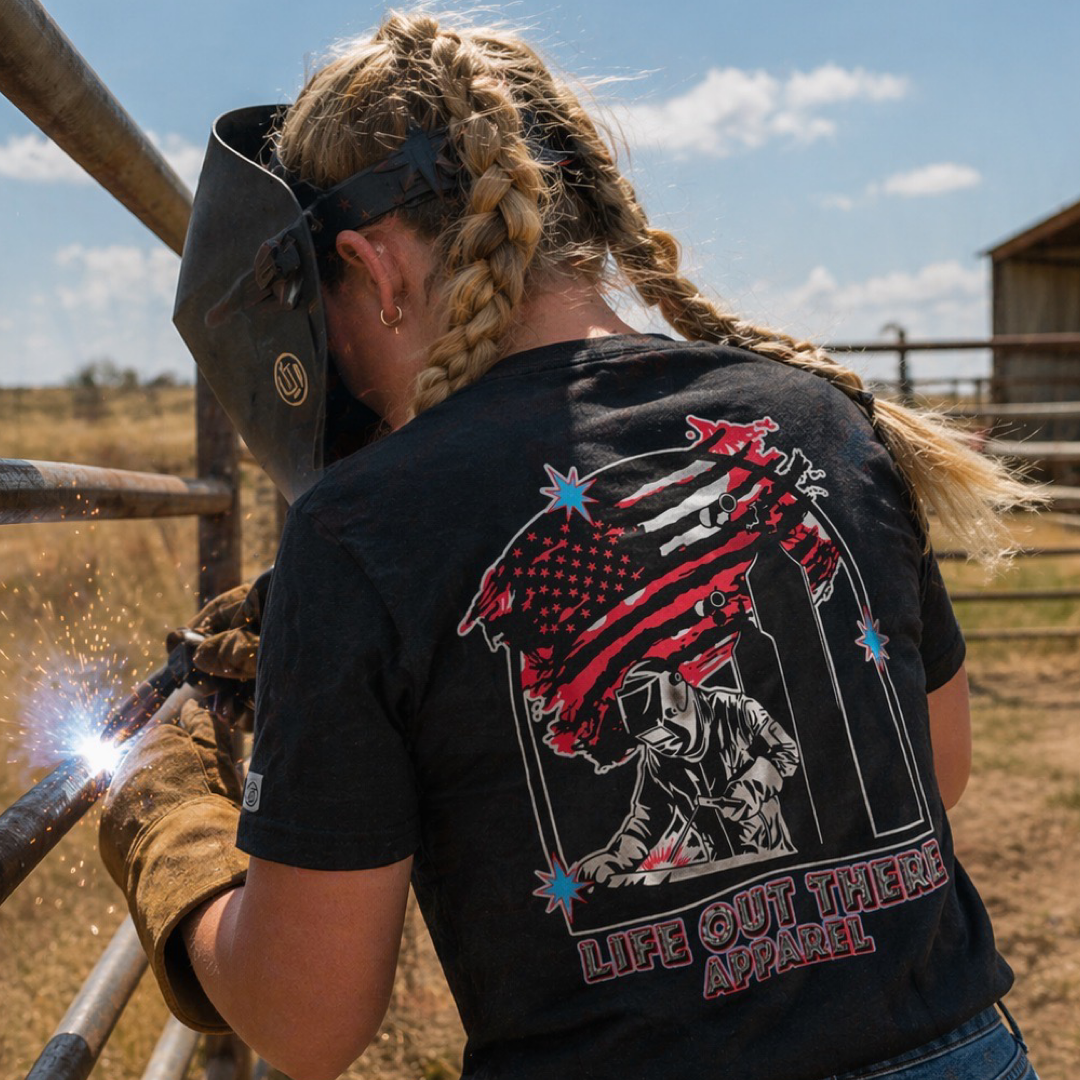 A woman with braided hair welds a metal fence outdoors, wearing the Tack It Tee - Black by Life Out There Apparel. The unisex T-shirt features an American flag, a welder, and bold “Life Out There Apparel” text—ideal for proud welders.